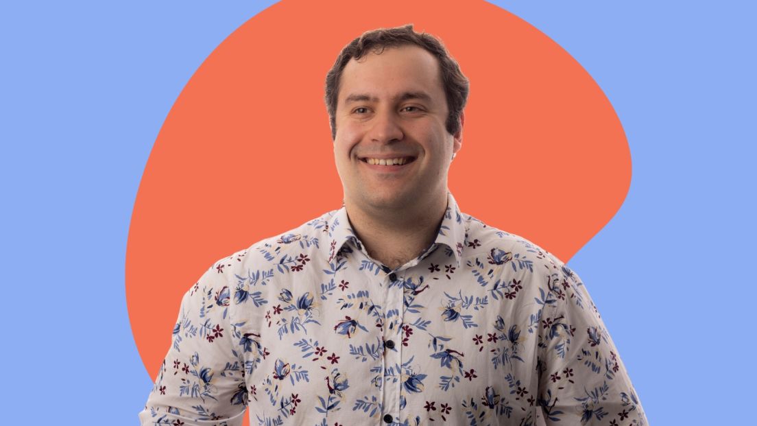 A smiling man with short dark hair, identified as Michael Theo, is wearing a white button-up shirt with a blue and maroon floral pattern. He is posed against a colorful background featuring a light blue backdrop with a large orange abstract shape behind him.