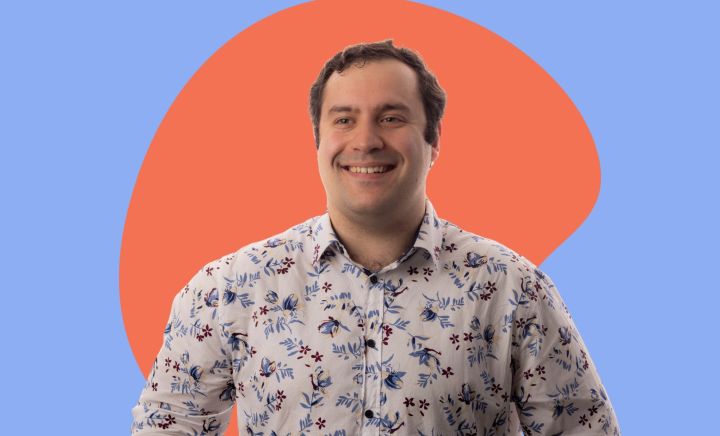 A smiling man with short dark hair, identified as Michael Theo, is wearing a white button-up shirt with a blue and maroon floral pattern. He is posed against a colorful background featuring a light blue backdrop with a large orange abstract shape behind him.