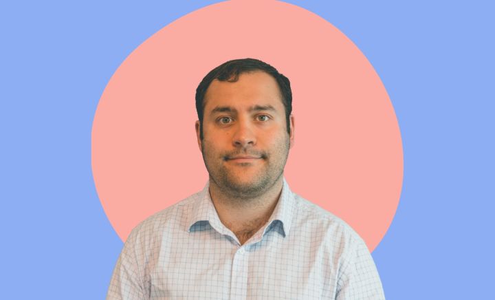 A man named Michael Theo with short, dark brown hair and light stubble is shown against a colorful background. He is wearing a light-colored, checkered button-down shirt. The backdrop features a large pink circle over a blue background, creating a simple, modern look. Michael faces the camera with a neutral expression.