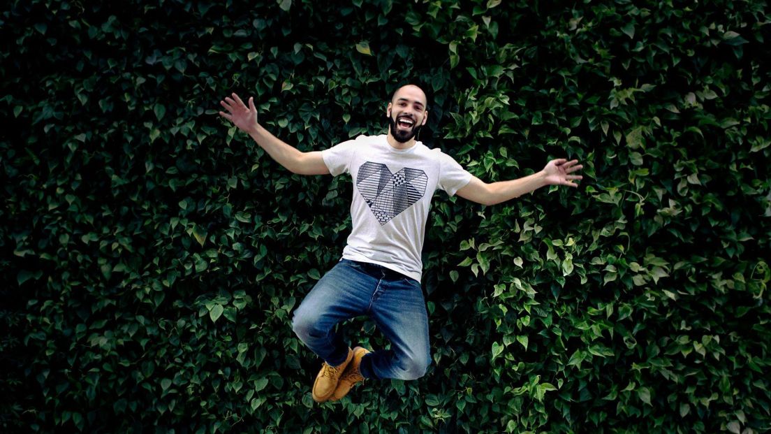 A man is mid-jump in front of a vibrant green wall covered in dense, leafy plants. He has a joyful expression, arms spread wide, and is wearing a white T-shirt with a geometric heart design, blue jeans, and tan boots. The floor beneath him is light gray tile, contrasting with the lush greenery behind him.