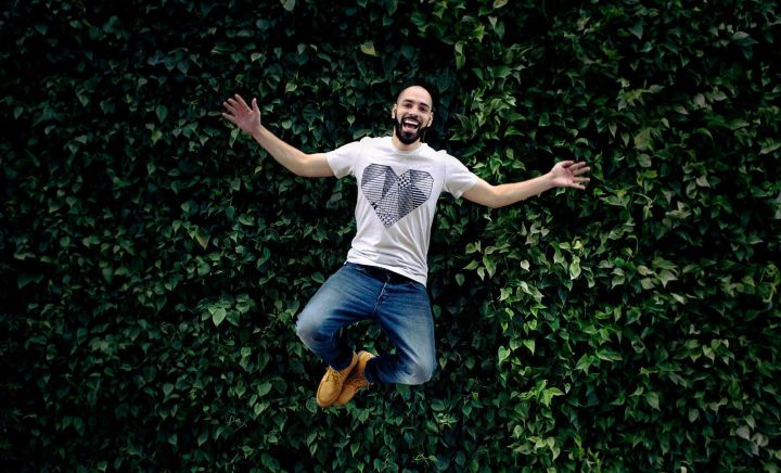 A man is mid-jump in front of a vibrant green wall covered in dense, leafy plants. He has a joyful expression, arms spread wide, and is wearing a white T-shirt with a geometric heart design, blue jeans, and tan boots. The floor beneath him is light gray tile, contrasting with the lush greenery behind him.