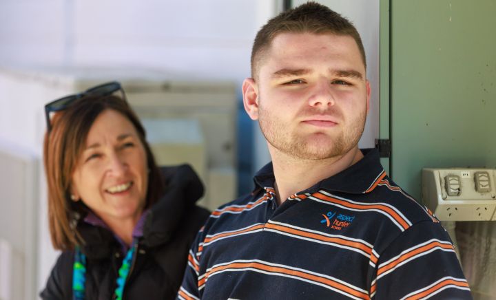 A young man with short hair and a trimmed beard stands outside, looking off into the distance with a thoughtful expression. He is wearing a navy blue, orange, and white striped polo shirt with a logo that reads "Aspect Hunter School." Behind him, a woman with shoulder-length brown hair smiles warmly in his direction. She is dressed in a black jacket and has sunglasses resting on her head. The setting appears to be outdoors, with soft, natural lighting and a relaxed atmosphere.